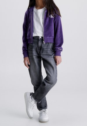 Sudadera morada con cremallera, camiseta blanca debajo, jeans grises, zapatillas blancas. La sudadera tiene un logo y puños acanalados. Tejido suave, corte casual.
