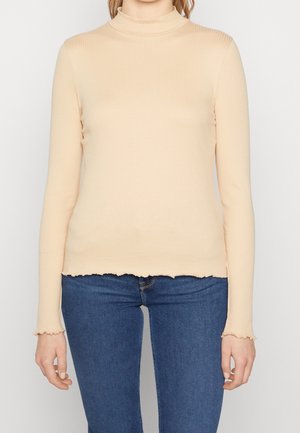 Žena v béžovém žebrovaném roláku s dlouhým rukávem a vroubkovaným lemem, v modrých džínách, stojící před jednoduchým pozadím.