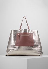 Metallinhohtoinen hopeanvärinen kangaskassi, jossa on teksturoitu pinta, vaaleanpunainen nahkainen etutasku ja kaksinkertaiset kantokahvat.