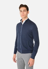 Cardigan zip con zip di colore navy realizzato in materiale a maglia fine, dotato di colletto alto, due tasche frontali e polsini e orlo a costine.