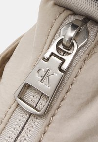 Tejido beige con una textura suave, con un tirador de cremallera plateado que muestra el logo "CK". La cremallera está centrada a lo largo de la costura.