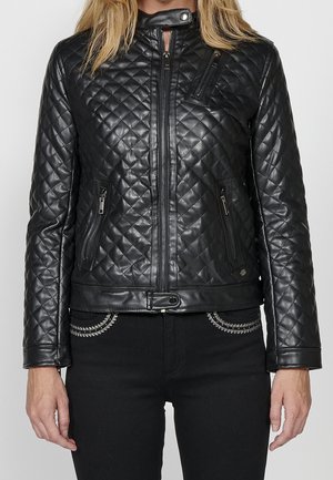 Femme portant une veste en cuir matelassée noire avec des poches zippées, un col pressionné, et un détail ceinture à la taille, associée à un pantalon noir avec une bordure argentée.