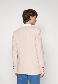 Helles rosa Blazer mit strukturiertem Design, Schlitze hinten und goldfarbene Knöpfe an den Ärmeln. Textiles Gewebe, taillierter Schnitt.