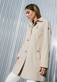 Beige Trenchcoat aus leichtem Material, mit Kragen, zwei Knöpfen, seitlichen Taschen und einem schlanken Design.