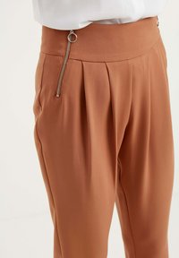 Bruine geplooide broek met een gladde textuur, voorzien van een zijritssluiting en een hoge tailleband, die de op maat gemaakte pasvorm benadrukt.