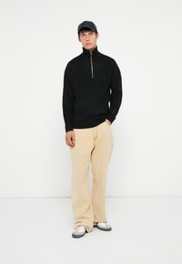 Pull noir à col zippé, pantalon beige à jambes larges et chaussures grises. Le pull possède des poignets et un ourlet côtelés, tandis que le pantalon a une texture lisse.