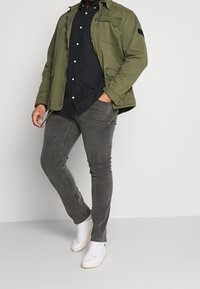 Olivgrön jacka med knäppning och bröstficka, svart skjorta med knappar, grå slim-fit byxor och vita sneakers.