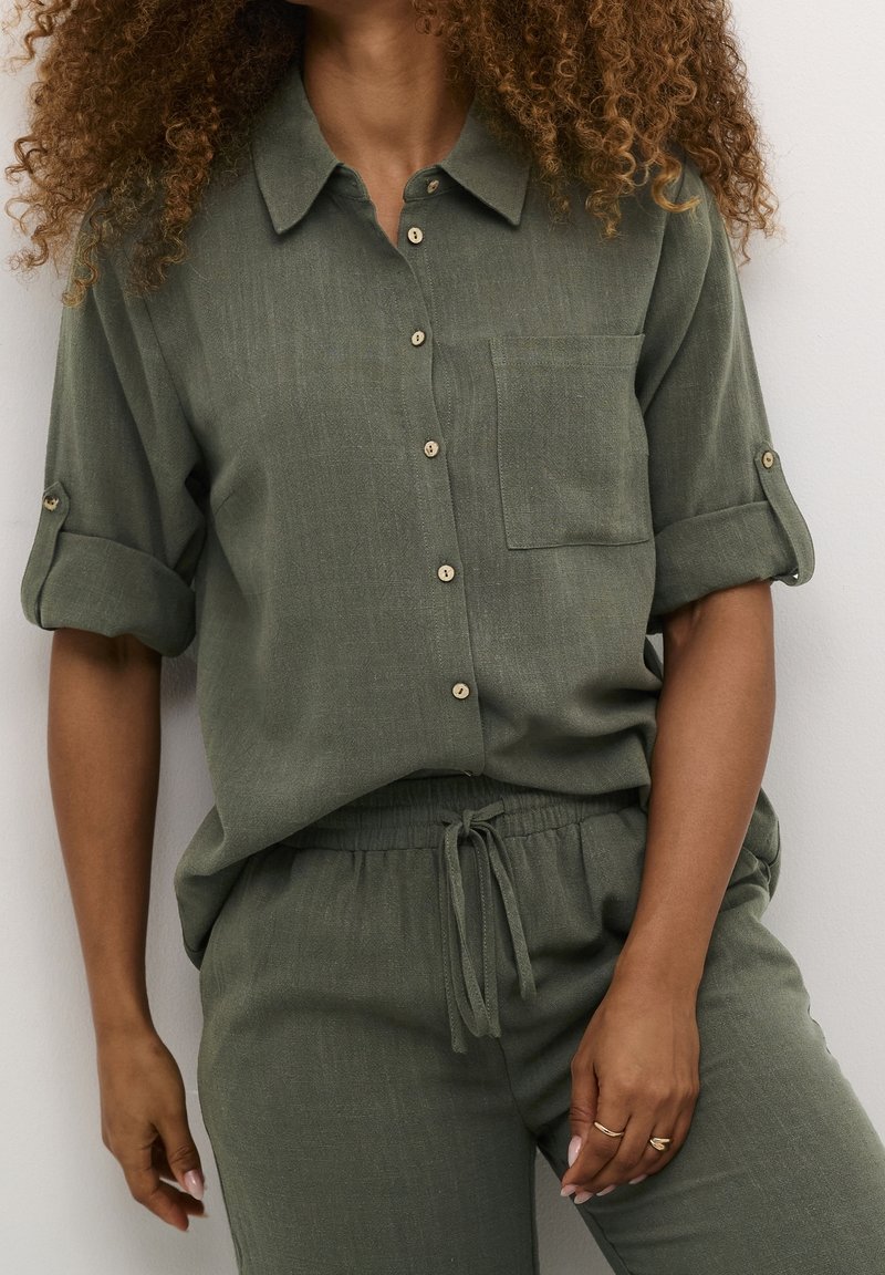 Donna che indossa una camicia verde oliva con bottoni e maniche arrotolate, abbinata a pantaloni con coulisse dello stesso colore, in piedi contro un muro bianco.