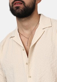 Hellbeige, strukturierte Button-Up-Bluse mit weichem Kragen und einer dekorativen Anhängerkette mit einem vertikalen Balken.
