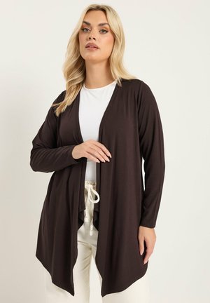 Blonde vrouw met een zwarte open cardigan over een wit topje en crèmekleurige broek met trekkoord, poserend tegen een effen lichte achtergrond.