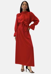 Robe longue en satin rouge avec manches longues, col haut, et ceinture nouée à la taille. Le tissu a une texture lisse et brillante. Fente sur un côté.
