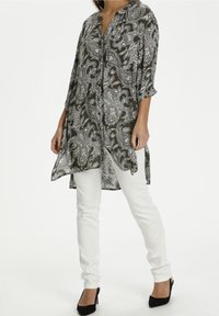 Tunique longue et ample de couleur vert foncé avec un motif cachemire blanc ; associée à un pantalon blanc ajusté et des talons pointus noirs.