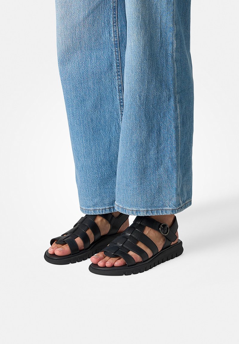 Personne portant un jean large bleu clair et des sandales en cuir noir avec plusieurs brides et une boucle, debout sur une surface blanche.