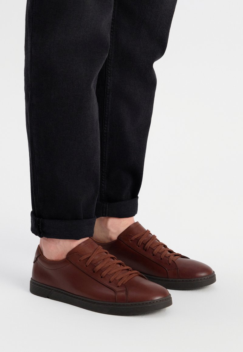 Brązowe skórzane trampki z płaskimi sznurowadłami i niskim profilem. Gumowa podeszwa z teksturowanym gripem i zaokrąglonym czubkiem. W połączeniu z ciemnymi, podwiniętymi dżinsami.