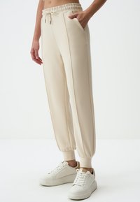 Beige joggings met een relaxte pasvorm, elastische tailleband, trekkoorddetail en zijzakken, gecombineerd met witte sneakers.
