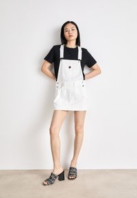 Bela denimova suknja na bretele z z adjustable straps, sprednja žepa in srebrni gumbi, kombinirana s črnimi natikači z odprtim prstom in črno majico.