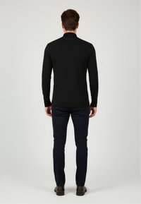 Langärmliges schwarzes Polohemd mit hohem Kragen, aus glattem Stoff, mit einem Logo am Nacken; kombiniert mit navyfarbigen Hosen und braunen Schuhen.