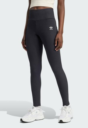Leggings negros de tiro alto con textura acanalada, que presentan un pequeño logo blanco de Adidas en la cadera izquierda. Combinados con zapatillas deportivas blancas.