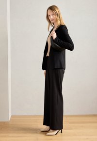Blazer noir cintré sur un top beige, associé à un pantalon noir à jambes larges et des escarpins crème à bout pointu. La mannequin tient un sac à main noir.
