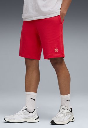 Mężczyzna w czerwonych spodenkach Puma, białych skarpetkach Puma i białych butach sportowych Puma, stojący z jedną ręką w kieszeni na neutralnym tle.