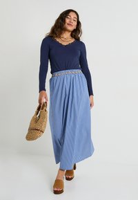 Top largo de manga en color azul marino con ribete de encaje, combinado con una falda maxi a rayas azules que presenta una cinturilla estampada. Sandalias de plataforma marrones y bolsa de paja.