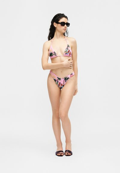 Femme portant un bikini rose à motifs floraux tropicaux, des sandales noires glissantes et des lunettes de soleil noires, posant debout contre un fond blanc.