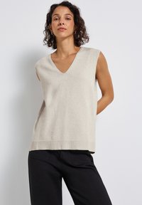 Kvinna med krulligt mörkt hår som bär en ärmlös beige V-krags jumper och svarta byxor, stående mot en ljus enfärgad bakgrund.