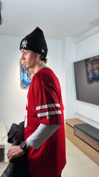 Ung person iført en rød sportstrøje og sort hue står indendørs og holder en sort jakke, med et TV og en elektrisk pejs i baggrunden.