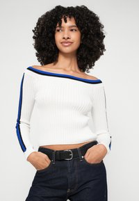 Fehér bordás, vállon kívül viselhető pulóver kék és fekete csíkos díszítéssel az ujjain, amelyet magas derekú sötét farmernadrággal és fekete övvel párosítanak.