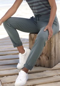 Grüne Jogginghosen mit umgeschlagenen Säumen, aus weichem Material, kombiniert mit weißen Sneakers. Streifenhemd im Hintergrund sichtbar.