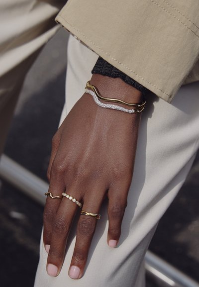 Bracciali d'oro e d'argento su una mano, con un design curvo e dettagli in cristallo. Vari anelli, compresi uno attorcigliato e uno a perline.