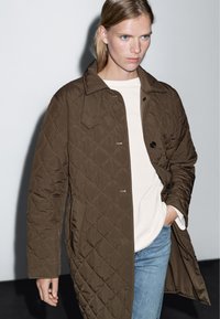 Hnědý prošívaný kabát s límcem a zapínáním na knoflíky, s bočními kapsami a volným střihem, kombinovaný se světle barevným topem a džíny.