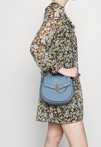 Borsa a tracolla in pelle blu con forma curva, dotata di chiusura a patta frontale, dettaglio con catena dorata e superficie testurizzata, indossata con un vestito floreale.