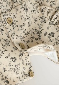Tissu beige à motif floral avec de petites fleurs noires, comportant deux boutons ronds beiges, un détail froncé, et une étiquette de vêtement.