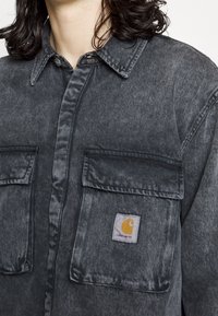 Tmavě šedá džínsová košile se zapínáním na knoflíky, dvěma náprsními kapsami a žlutou nášivkou na levé kapse. Měkká textura.