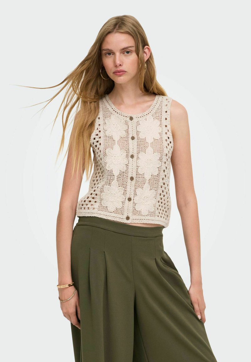 Jeune femme aux longs cheveux blonds portant un haut beige sans manches en crochet avec des motifs floraux et un pantalon large vert olive.