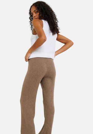 Femme avec des cheveux bouclés regarde par-dessus son épaule, portant un haut sans manches blanc et un pantalon beige côtelé taille haute, le tout sur fond blanc.