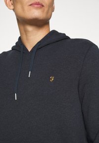 Haut à capuche gris en tissu doux avec un cordon de serrage. Présente un petit logo jaune sur la poitrine gauche et des accents en argent sur les cordons.