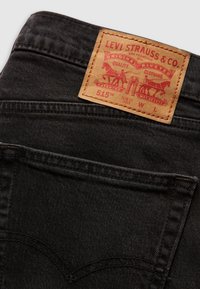 Poche arrière d'un jean en denim noir avec un patch en cuir couleur beige de Levi Strauss & Co. montrant deux chevaux et un texte incluant "Original Riveted" et "515 Slim Fit Taper."