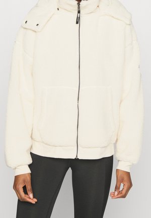 Personne portant une veste polaire blanche zippée avec un col montant et de grandes poches avant, associée à des leggings noirs.