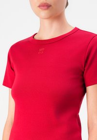 Woman wearing a fitted red short-sleeve top with "HUGO" embroidered at the center near the neckline.