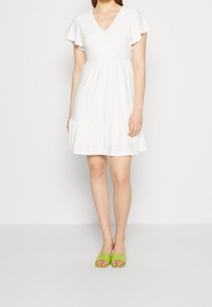 Femme portant une robe blanche à manches courtes avec un ourlet à volants et des sandales vertes à bouts ouverts, debout devant un fond clair uni.