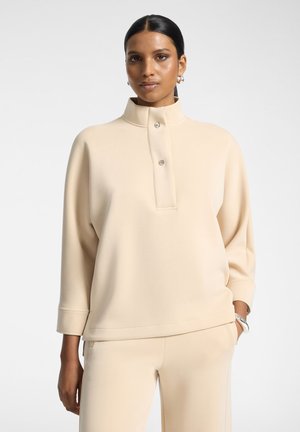 Femme aux cheveux foncés portant un haut beige à manches longues avec des boutons et un pantalon assorti, debout avec une main dans la poche sur un fond uni.