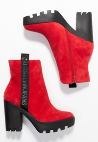 Botines de ante rojo con gruesos tacones y suelas negras, cremallera lateral y tira de tela negra etiquetada "Calvin Klein Jeans."