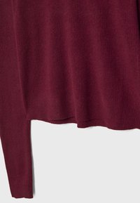 Pull en tricot bordeaux à manches longues, texture côtelée, avec un ourlet légèrement irrégulier sur un fond clair uni.
