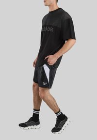 Hombre con pantalones cortos negros Reebok con paneles laterales blancos, camiseta negra Reebok a juego, calcetines negros con rayas blancas y zapatos deportivos negros.