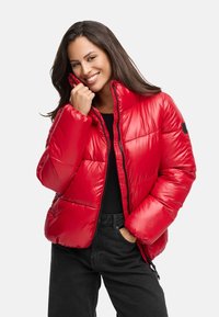 Chaqueta acolchada roja con cuello alto, acabado texturizado y cremallera frontal. Presenta acentos negros y se lleva sobre una camiseta negra y pantalones oscuros.