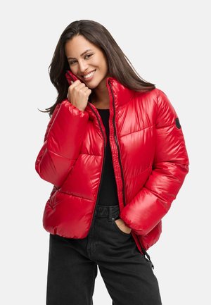 Giacca a vento rossa con colletto alto, finitura texturizzata e zip anteriore. Presenta dettagli neri ed è indossata sopra una maglia nera e pantaloni scuri.