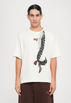 Cream short-sleeve t-shirt featuring a black braided hair illustration with white flowers and brown accents. Casual relaxed fit.