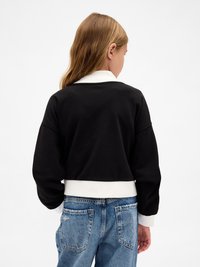 Giovane con lunghi capelli castano chiaro che indossa un maglione bianco e nero e jeans in denim blu, voltato di spalle contro uno sfondo bianco.